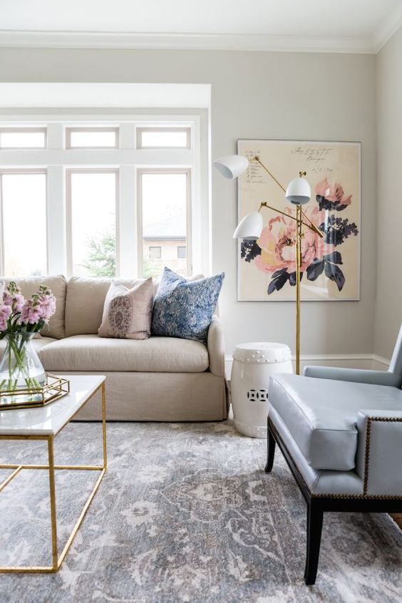 a neutral living room with grey walls, a tan sofa and a blue leather chair, a floral rug, floral artwork and some fresh blooms