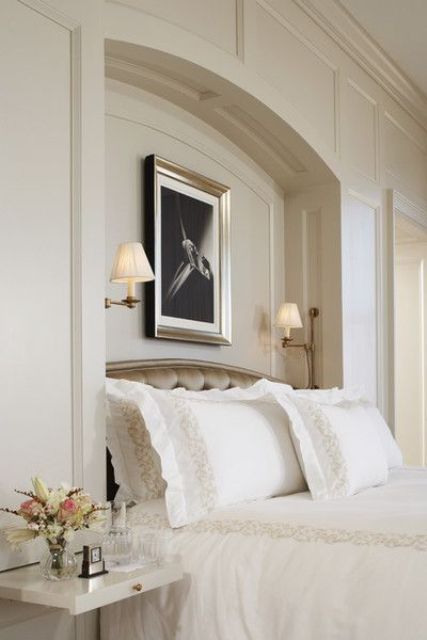 a sophisticated tan bedroom with an arched niche with sconces and art, with an upholstered bed with neutral bedding