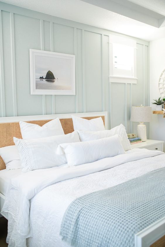 a beautiful coastal bedroom with a pale green accent wall, a bed with a rattan headboard, neutral bedding and some art