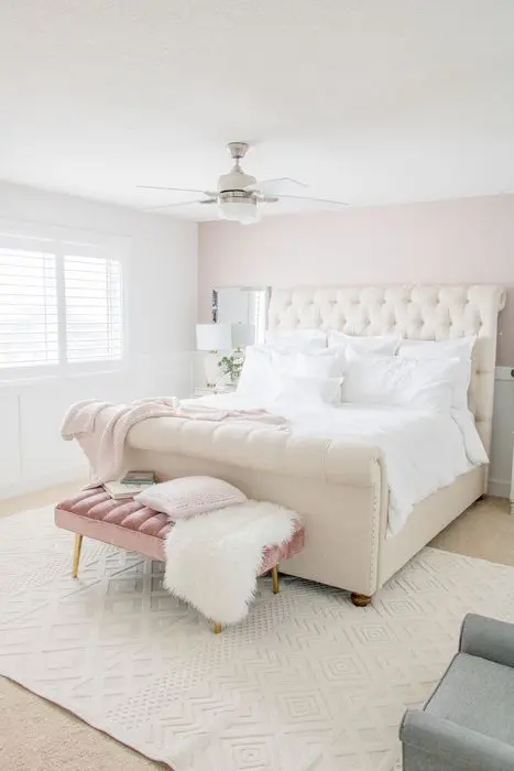 a bedroom with a pale pink accent wall, a creamy upholstered bed, a dusty pink upholstered bench and a grey chair