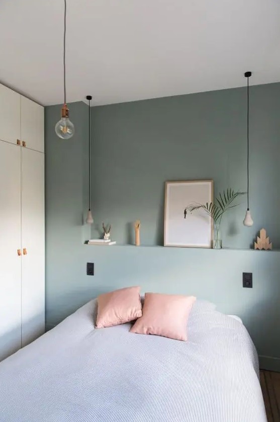 a bedroom with a pastel green accent wall, a bed with grey and pink bedding, a white wardrobe and pendant bulbs
