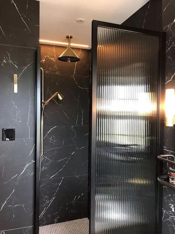 a black bathroom clad with marble, with a fluted glass space divider, brass touches for a more elegant and chic look