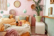 a boho pastel bedroom with a pink accent wall and green walls, a bed with muted color bedding, a macrame lamp and pastel pillows