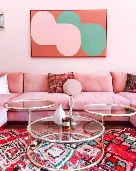 a bright and feminine living room with light pink walls, a pink sofa, a glass coffee table, a bold artwork and a colorful rug with prints