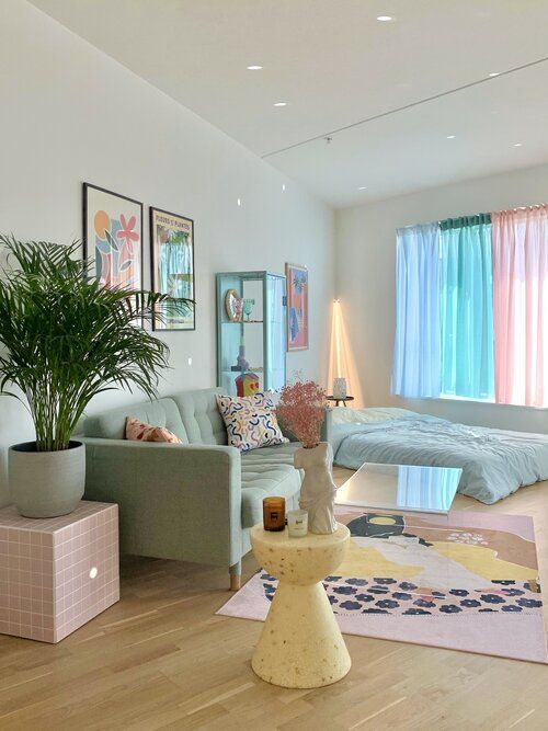 a candy-colored living room with a mint sofa and a bed, pastel curtains, a pink side table, a yellow side table, a pastel printed rug and bold artwork