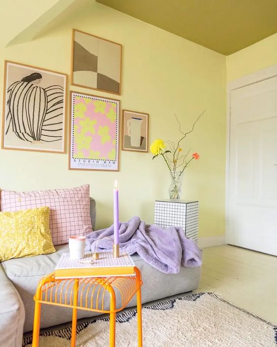 a candy-colored living room with light yellow walls, a low grey sofa with pastel pillows, a gallery wall, an orange stool and a lilac candle