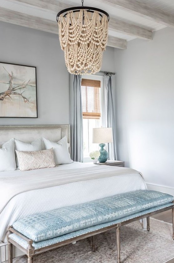 a coastal bedroom in light and pastel blues, with a chic bed and bench, a wood bead chandelier, blue textiles and an artwork