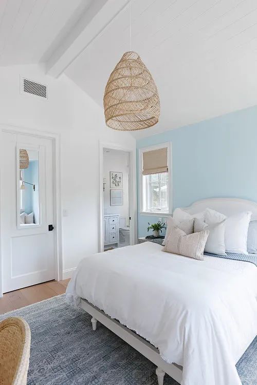 a coastal bedroom with a pastel blue accent wall, a neutral bed with neutral bedding, a woven pendant lamp and a woven chair