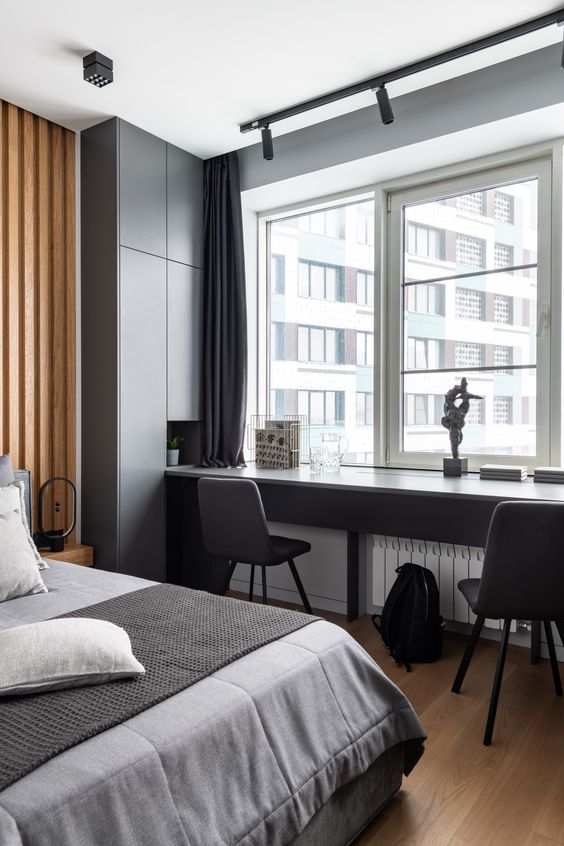 a contemporary bedroom in graphite grey, a slatted wood accent wall, a bed with grey bedding, a windowsill desk and dark chairs