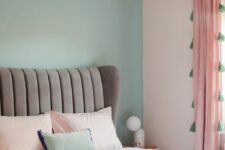 a cool pastel bedroom with a mint blue accent wall, a grey upholstered bed, pastel and coral bedding and pink curtains