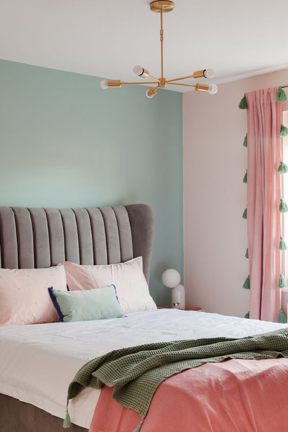 a cool pastel bedroom with a mint blue accent wall, a grey upholstered bed, pastel and coral bedding and pink curtains