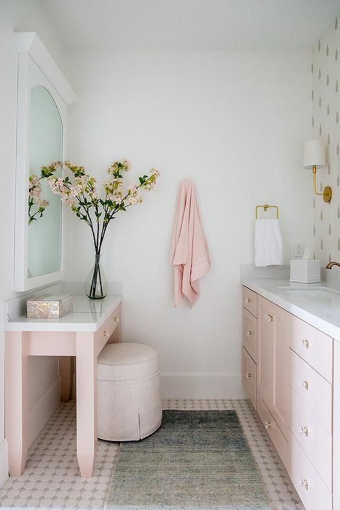 a cute pink bathroom with pink cabinetry and a vanity, a blush pouf and wallpaper, a mirror cabinet is very girlish