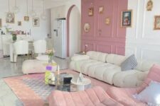 a delicate living room with a color block pink and white wall, a creamy low sofa, pink ottomans, neutral tables