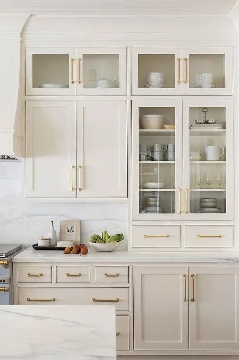 a delicate tan kitchen with shaker and glass front cabinets, gold handles and a white marble backsplash and countertops