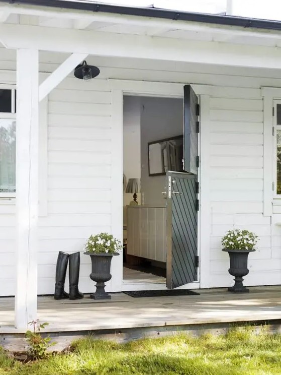 a farmhouse entrance with a black diagonal stripe Dutch door, black urns with white blooms and greenery and riding boots is very cozy and bold