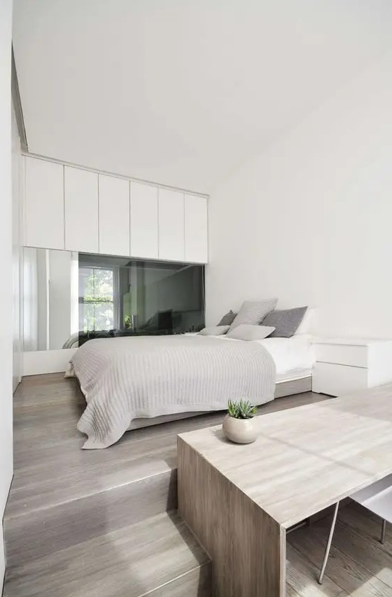 a functional minimalist bedroom with sleek white cabinets, a glass wall, a bed with neutral bedding, a nightstand and a desk plus a chair by the window