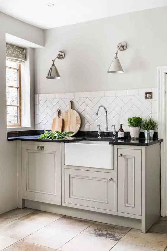 a greige vintage kitchen with cool cabinets, black stone countertops, a white herringbone tile backsplash and metal sconces