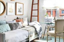 a light-filled bedroom with neutral walls, a convertible sofa, a glass trestle desk, a neutral chair and a bold gallery wall plus a ladder