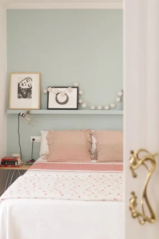 a light green bedroom with a bed with pink and white bedding, an open shelf and a nightstand plus black and white artwork