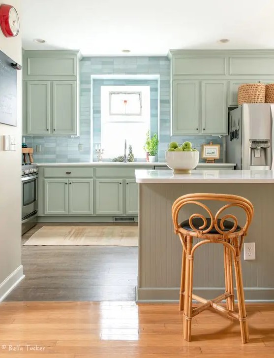 a lovely sage green kitchen with shaker cabinets, a blue skinny tile backsplash a slatted kitchen island and vintage rattan stools