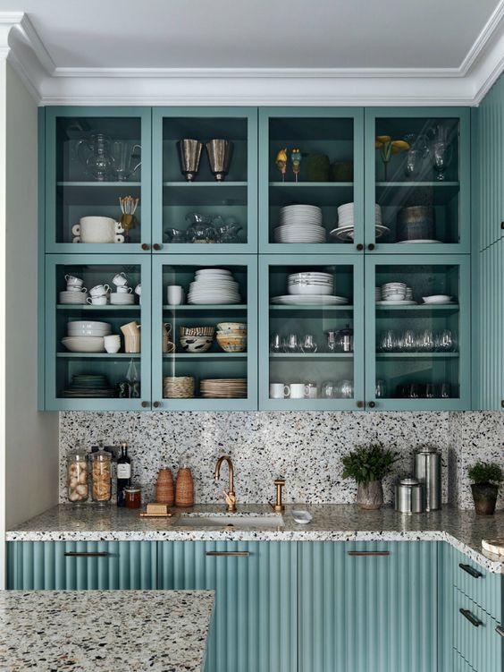 a lovely turquoise kitchen with fluted and front glass cabinets, a white terrazzo backsplash and countertops and brass handles
