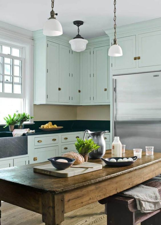 a mint green farmhouse kitchen black granite countertops and a backsplash, brass handles, a stained rough wood table and lamps on chain
