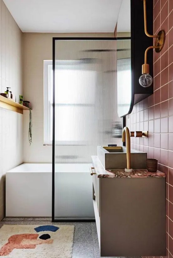 a modern bathroom with dusty pink tiles and grey ones, a graphic rug, a bathtub and a reeded glass space divider and a large mirror