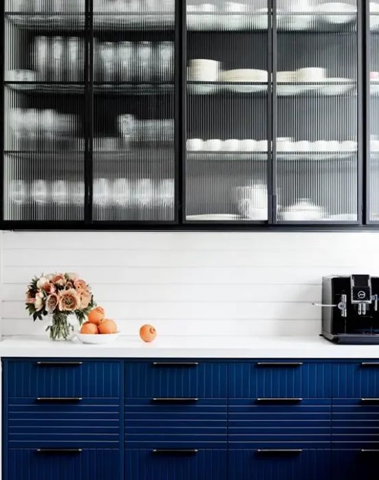 a modern contrasting kitchen with fluted glass and navy fluted cabinets, a white beadboard backsplash and white stone countertops