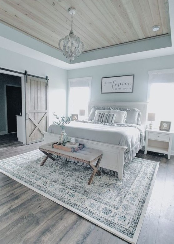 a modern farmhouse bedroom in pastel blue shades with white furniture, an upholstered bench, a vintage chandelier and ruffle bedding