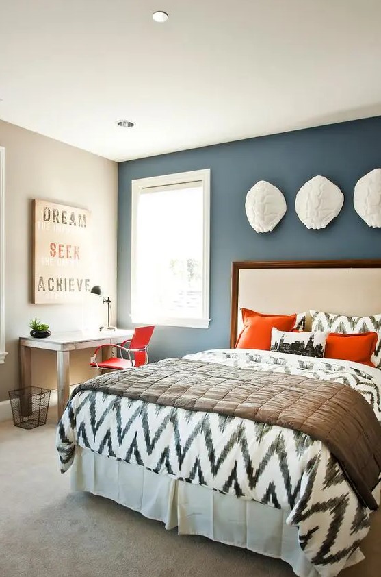 a modern farmhouse bedroom with a navy accent wall, an upholstered bed, printed bedding and bright pillows, a stained desk and an orange chair