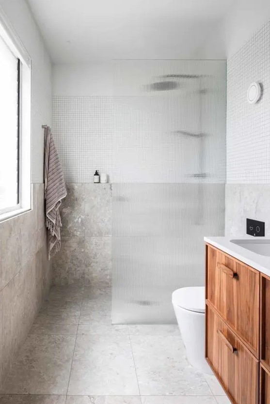 a modern laconic bathroom with neutral stone tiles, a shower space with a fluted glass space divider, a floating vanity and a window with much light