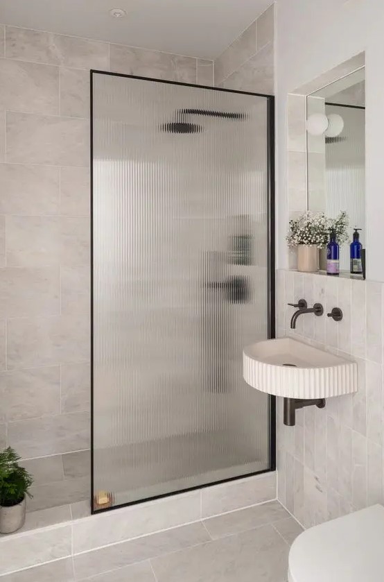 a neutral bathroom with stone tiles, a shower space with a fluted glass space divider, a wall-mounted sink and a mirror with a shelf