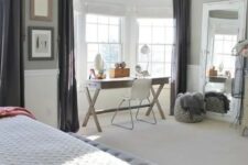 a neutral farmhouse bedroom with grey and white paneled walls, a large bed and a grey upholstered bench, a trestle desk and a neutral chair in the bay window space