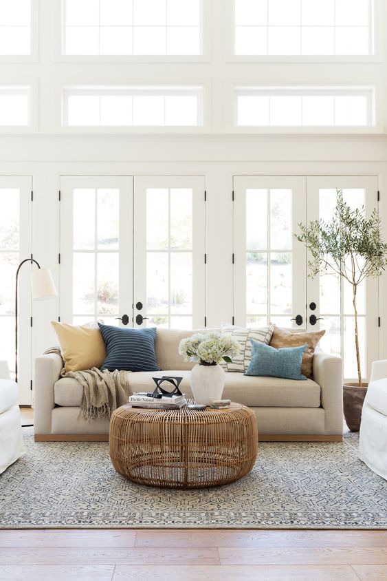 a neutral farmhouse living room with white French doors, a neutral sofa and printed pillows, a rattan table and potted plants