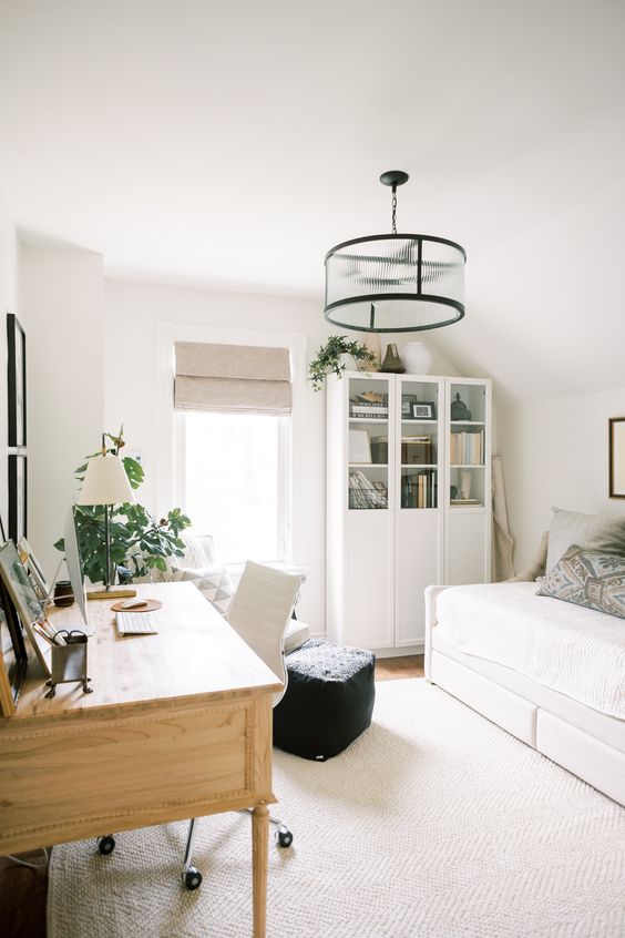 a neutral guest bedroom with a white bed and bedding, a bookcase, a stained desk, a white chair and a black pouf