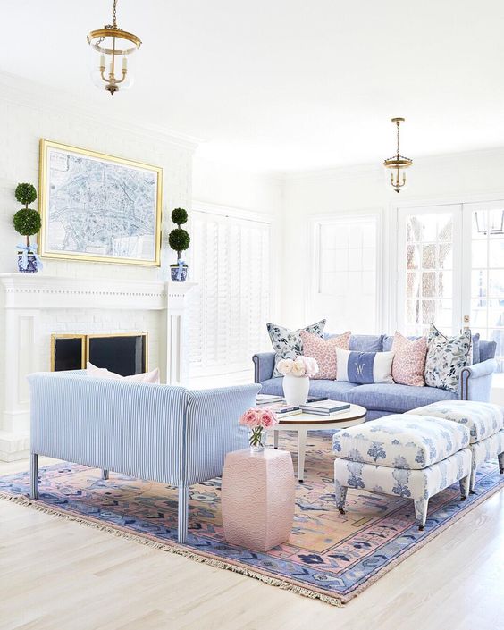 a neutral living room with a fireplace, periwinkle sofas, printed ottomans, a pastel printed rug, a round table and a small pink side one