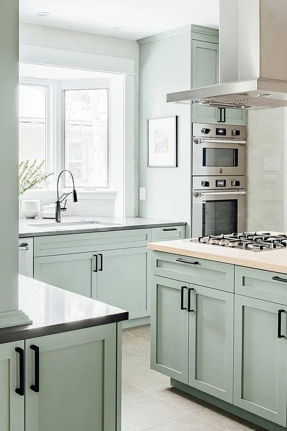 a pale mint green kitchen with shaker cabinets, grey granite and butcherblock countertops, black handles and stainless steel appliances