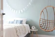 a pastel blue bedroom with a grey bed and neutral bedding, a rattan pendant chair, a small stool as a nightstand and a woven rug