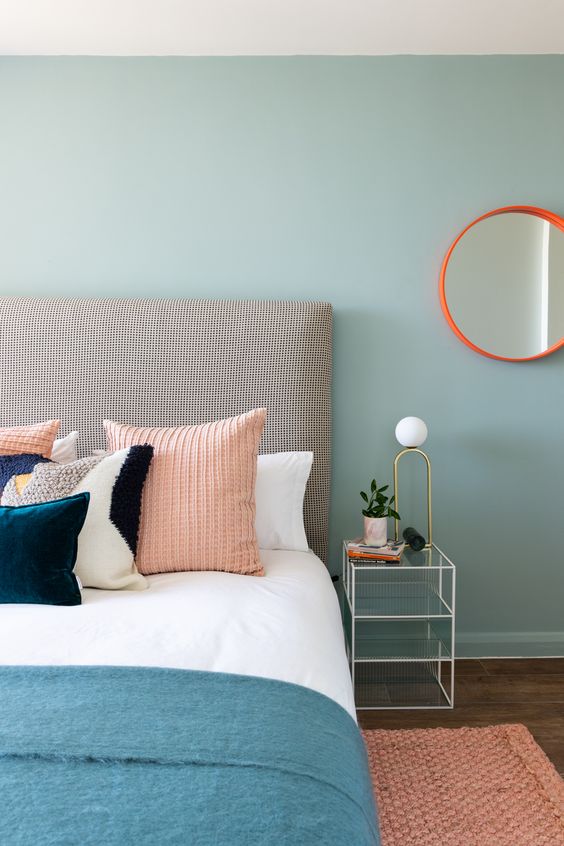 a pastel blue bedroom with an upholstered bed and pastel bedding, a pink rug and a mirror in an orange frame