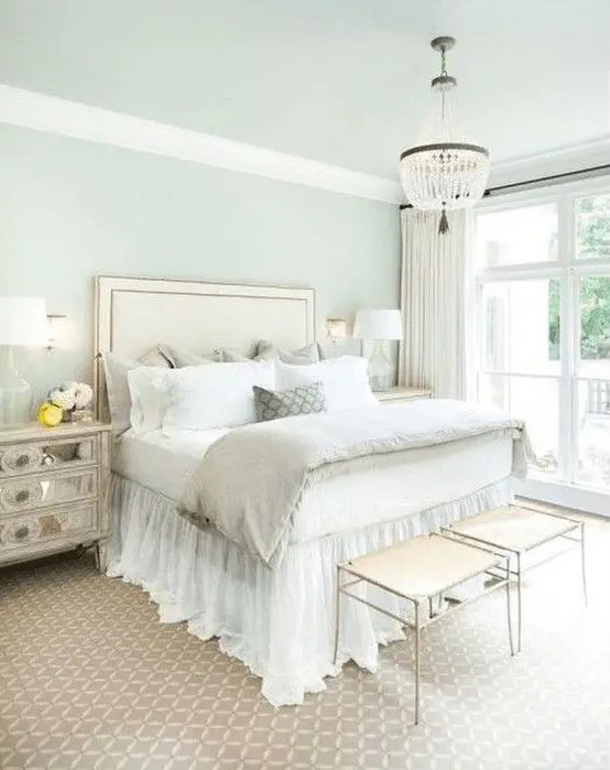 a pastel glam bedroom with an upholstered bed, a crystal chandelier, mirror nightstands and elegant stools
