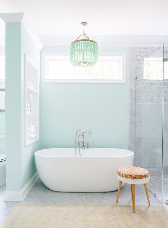 a pastel green bathroom with a glass-enclosed shower space, an oval tub, a niche with shelves and a green bead chandelier