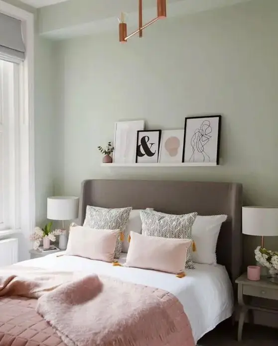 a pastel green bedroom with an upholstered bed, pink bedding, a ledge with artworks and a chandelier