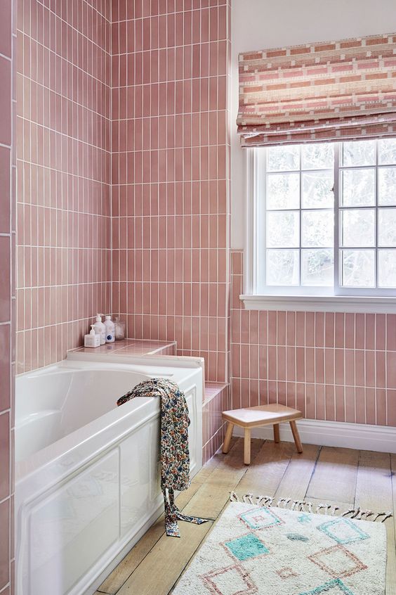 a pastel pink bathroom with skinny tiles, a bathtub and several built-in shelves, a blush curtain and a boho rug