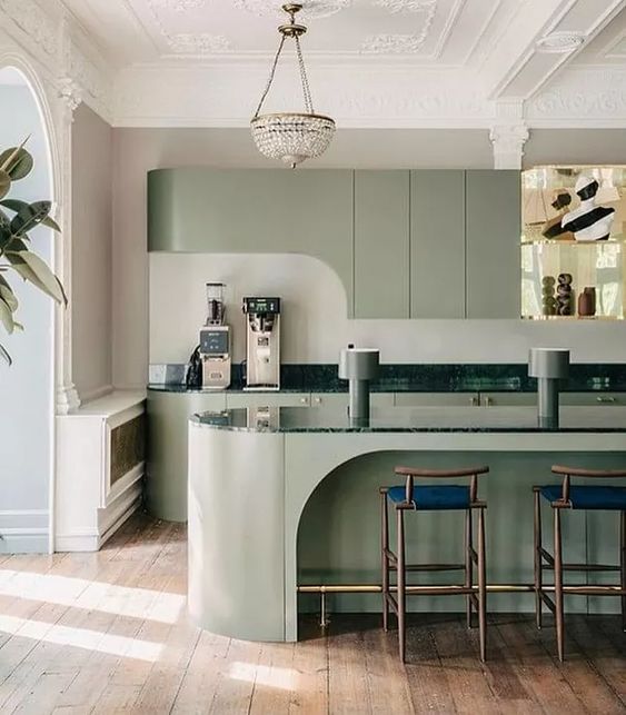 a plain green kitchen with sleek cabinets, a curved kitchen island with dark green stone countertops, elegant stools and a chic chandelier