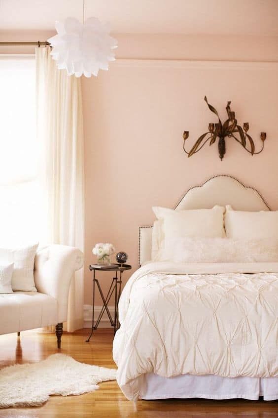 a pretty pastel pink bedroom with creamy upholstered furniture, neutral bedding, a white pendant lamp and darkened metal decor