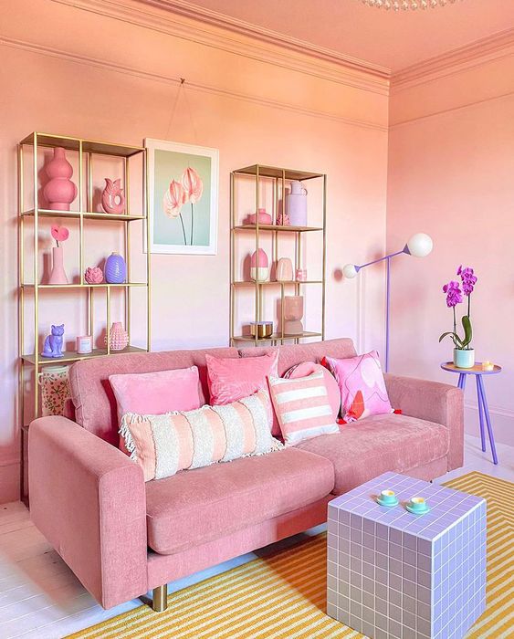 a pretty pink living room with gold shelving units, a pink sofa with pastel pillows, a lilac cube table and a yellow rug