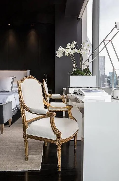 a refined black bedroom with an upholstered bed with neutral bedding, a grey upholstered bench, a white desk and a refined chair by the window