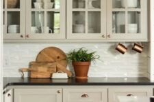 a refined greige kitchen with shaker and glass front cabinets, a white embossed tile backsplash and black countertops is a beautiful farmhouse solution