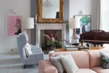 a refined neutral living room with a non-working fireplace, a mirror in a gilded frame, a low pink sofa, blue chairs, a piano and a crystal chandelier
