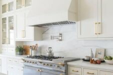 a refined vintage creamy kitchen with shaker cabinets, a white quartz backsplash and countertops, brass and gold fixtures